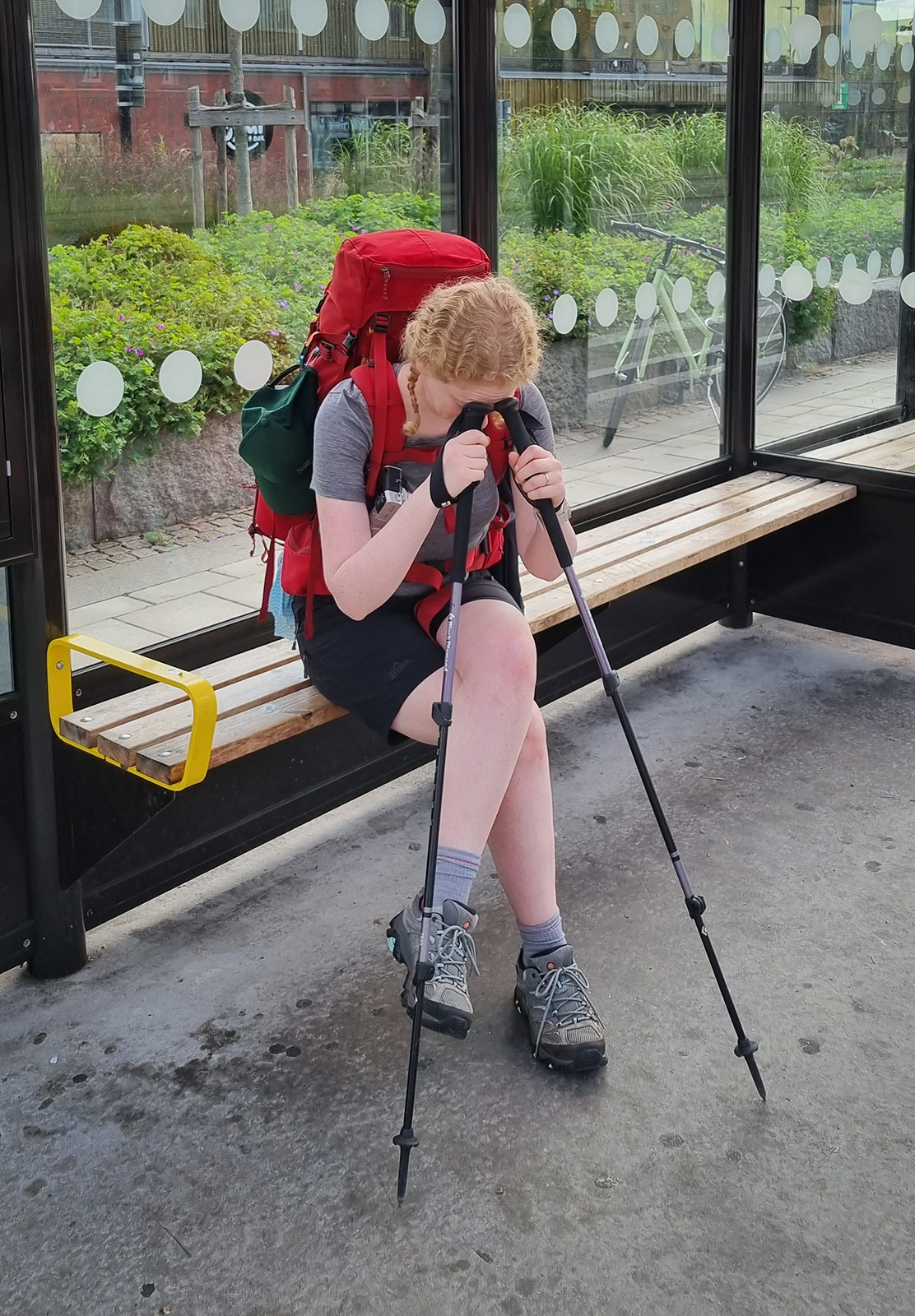 Woman sat in bus stop resting head on trekking poles